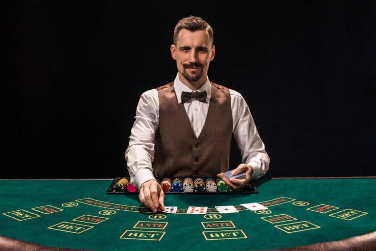 UK Live Casino Portrait of a croupier is holding playing cards, gambling chips on table. Black background. A young male croupier in a shirt, waistcoat and bow tie is waiting for you at the blackjack table